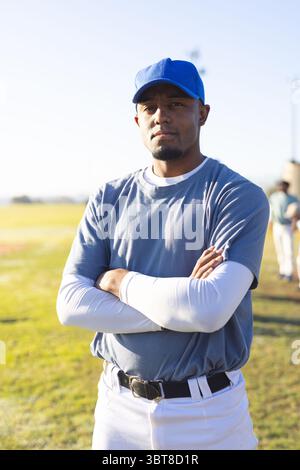African American male baseball player standing crossing arms on sunlit outfield wearing blue cap Stock Photo