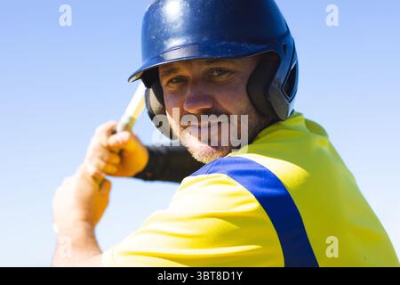 Male baseball batter wearing blue helmet and yellow jersey, swinging wooden bat on baseball field Stock Photo