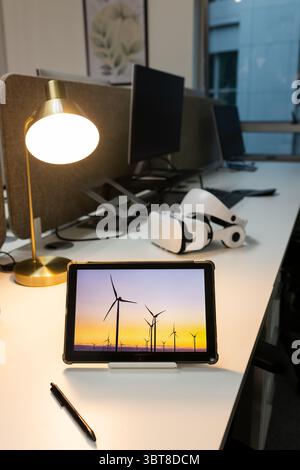 Tablet in protective case displaying wind turbines at sunset on desk with lamp and VR headset Stock Photo