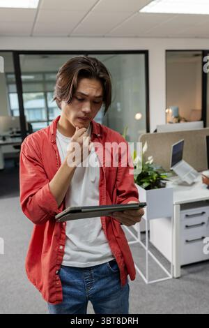 Asian man standing in modern office interior holding tablet and touching chin while studying screen Stock Photo