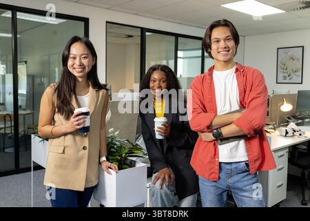 Diverse coworkers standing around planter in open office holding coffee and laptop Stock Photo
