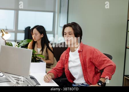 Asian coworkers working at modern office using silver laptop beside brass desk lamp and green plant Stock Photo