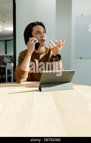 Asian man talking on smartphone and gesturing at wooden meeting table with tablet and whiteboard Stock Photo
