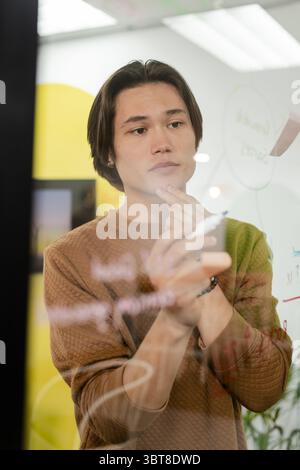 Asian man standing at glass whiteboard touching chin while drawing diagrams with marker in office Stock Photo