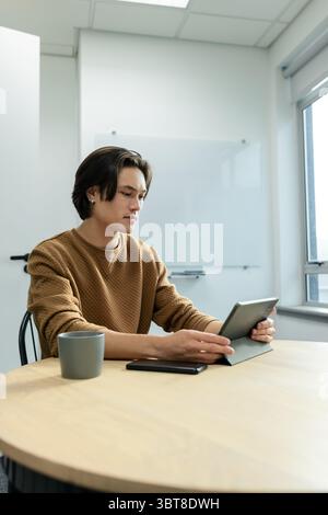 Asian man wearing brown sweater scrolling tablet at office with whiteboard smartphone and grey mug Stock Photo