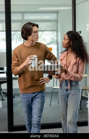 Diverse coworkers standing and talking while holding coffee cup and tablet in glass office Stock Photo