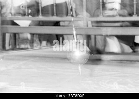 Bavo day in the Grote Kerk: test Foucault's pendulum Haarlem The Netherlands, Whizgle News, Dutch Desk, The Netherlands, 1950 - 2000 on 04-10-1991. These are the elements in the image. The scene captures a pendulum in motion, suspended from a slender wire. The pendulum itself is a shiny metal sphere, reflecting light in a subtle manner, creating a glimmer against a backdrop that suggests soft, muted tones. The surface beneath is textured, perhaps slightly uneven, hinting at a canvas or a work surface that captures intricate designs or patterns beneath it. In the background, there are hints of Stock Photo