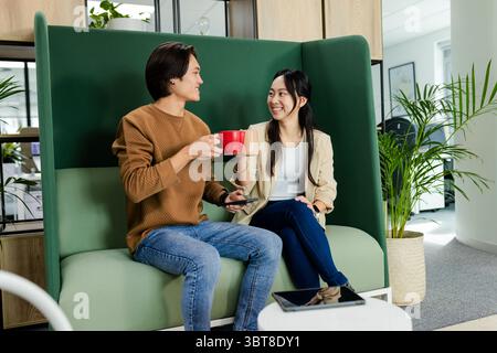 Asian coworkers toasting red and cream mugs on sofa in office breakout area with tablet Stock Photo