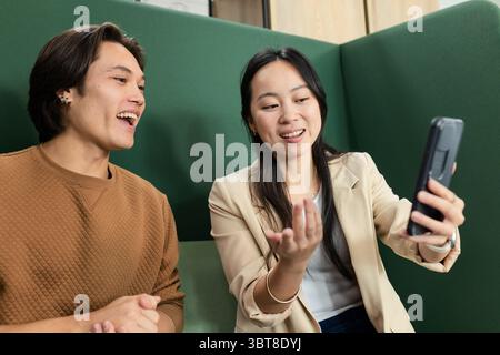 Asian colleagues sitting on green upholstered bench in office lounge, taking selfie with smartphone Stock Photo