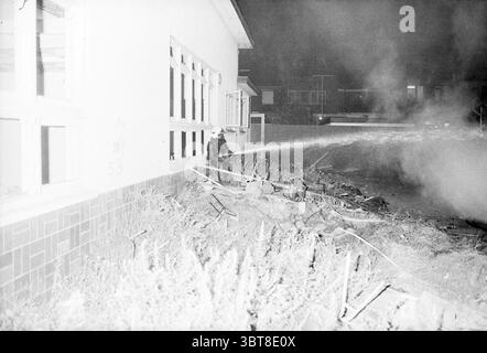 Fire on the IJmuiden municipal site IJmuiden The Netherlands, Whizgle News, Dutch Desk, The Netherlands, 1950 - 2000 on 28-08-1990. The image shows these topics. The scene captures a dramatic moment at night, illuminated by stark, bright light that creates a stark contrast with the surrounding darkness. In the foreground, a brave figure clad in firefighting gear is actively engaged in controlling a fire, directing a powerful stream of water towards the source of flames. The suit is mostly dark, making it stand out against the lighter background. To the left, there is a stark, white building, i Stock Photo