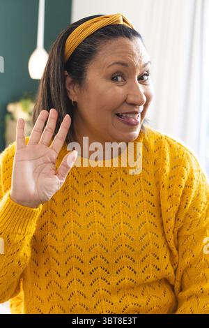 African American woman raising hand in greeting wearing yellow headband at home with pendant lamp Stock Photo