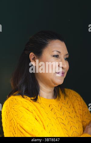 Mature woman posing in mustard yellow knitted sweater with silver nose ring against dark green wall Stock Photo