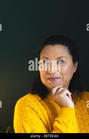 Middle-aged woman resting chin on hand against dark green wall wearing yellow sweater, copy space Stock Photo