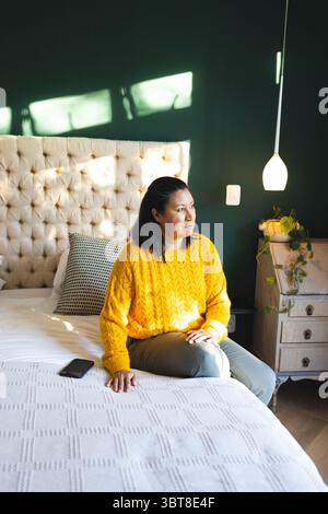Asian woman sitting on bed in mustard sweater, holding smartphone near nightstand plant, copy space Stock Photo
