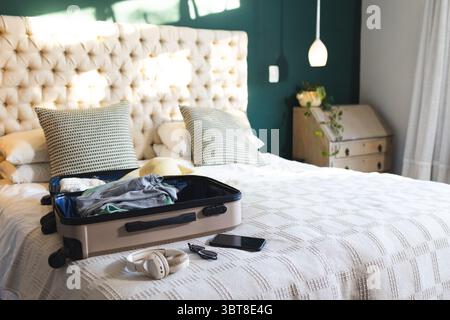 Open beige suitcase resting on made bed with patterned pillows and headphones against teal wall Stock Photo