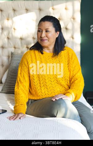 Woman sitting on bed with decorative pillows at home wearing mustard yellow sweater with smartphone Stock Photo