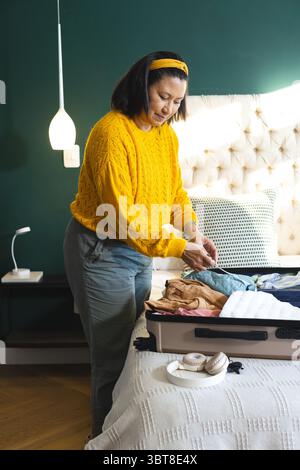 Woman sorting and folding clothes from open suitcase on bed in bedroom with dark green wall Stock Photo