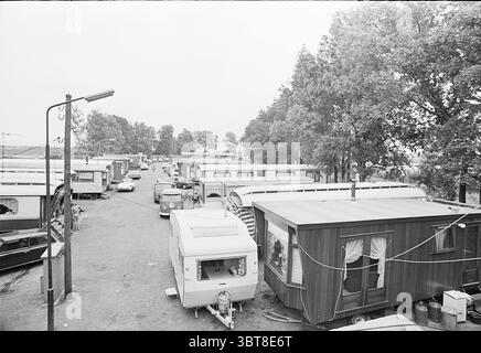Overview of a gypsy camp in the Waarderpolder Gypsies, Whizgle News, Dutch Desk, The Netherlands, 1950 - 2000 on 09-07-1974. The image includes these topics. The scene presents a view of a caravan park, characterized by an array of parked trailers and mobile homes lined along a narrow road. The atmosphere feels somewhat muted and nostalgic, dominated by a monochromatic palette that emphasizes the shades of gray. In the foreground, several caravans exhibit different architectural styles, some with distinctive overhangs and shutters, while others are more simplistic. A few have small steps leadi Stock Photo