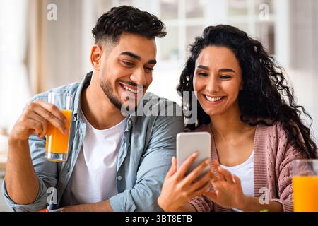 Portrait Of Cheerful Arab Lovers Using Smartphone During Breakfast In Kitchen Stock Photo