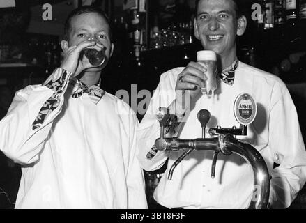 Dutch Beer Tapping Championship 2 men Hoofddorp The Netherlands, Whizgle News, Dutch Desk, The Netherlands, 1950 - 2000 on 09-01-1996. The image contains these topics. The scene captures two individuals standing behind a bar counter, both dressed in crisp white shirts complemented by patterned bow ties. Their attire gives off a polished yet approachable vibe. One person holds a pint glass filled with a dark beverage, while the other raises a glass of light-colored beer, both cheering with friendly smiles. The bar is well-stocked, with rows of bottles and glassware visible in the background, co Stock Photo