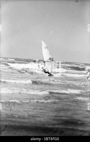 Paul Janssen Zandvoort Surfing Zandvoort, Whizgle News, Dutch Desk, The Netherlands, 1950 - 2000 on 29-06-1982. These topics are shown in the image. The scene captures a dynamic coastal environment, where the energy of the ocean meets the enthusiasm of water sports. In the foreground, a windsurfer skillfully navigates the waves, their body leaning intensely to maintain balance on the board. The sail, striking and billowing, features a distinctive pattern that draws the eye, blending with the vibrant blues and whites of the sea and surf. The board glides smoothly over the water, creating a slig Stock Photo