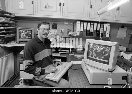 World Cup football competition on the computer through videotex Cruquius, Whizgle News, Dutch Desk, The Netherlands, 1950 - 2000 on 15-06-1990. These topics appear in the image. In a small, cozy workspace, a man sits at a wooden desk, immersed in his tasks. He has a neatly trimmed mustache and wears glasses, giving him a focused appearance. His attire features horizontal stripes of dark and light colors, lending a casual yet professional vibe. The desk is cluttered with papers, folders, and various office supplies, showcasing the busyness of his environment. To his side, two computer monitors Stock Photo