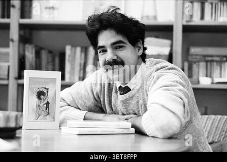 Writer Carlos M. Gase H'lem Haarlem Nederland, Whizgle News, Dutch Desk, The Netherlands, 1950 - 2000 on 23-01-1992. These topics are shown in the image. A young man with a thick, black mustache sits comfortably at a table, leaning slightly forward with a warm, welcoming smile. His hair is dark and voluminous, styled with a casual, slightly tousled look, reflecting a relaxed vibe. He wears a light sweater over a collared shirt, suggesting a blend of casual and thoughtful attire, suitable for a creative or scholarly setting. In front of him, there are a few books stacked neatly, one prominently Stock Photo