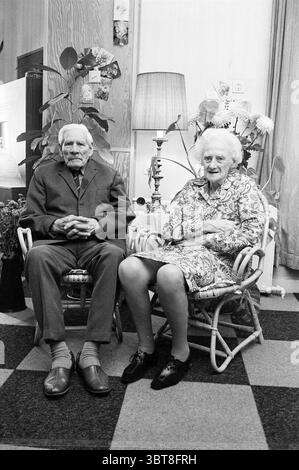 50-year-old couple De Bode Couples, Whizgle News, Dutch Desk, The Netherlands, 1950 - 2000 on 19-09-1972. These are the topics in the image. The scene depicts an elderly couple seated side by side in a cozy indoor setting. They are positioned in a pair of wicker chairs with a soft, textured fabric covering their seats. The man, dressed in a tailored gray suit, exudes a sense of dignity. His slicked-back hair is white, and he has a poised, thoughtful expression. He is wearing dark shoes complemented by light-patterned socks. Beside him, the woman is adorned in a vibrant floral-patterned dress, Stock Photo