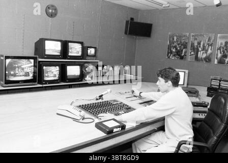 Jeroen Kosterman video editing Hillegom Video Hillegom, Whizgle News, Dutch Desk, The Netherlands, 1950 - 2000 on 09-12-1987. These are the topics in the image. The scene is set in a control room, characterized by a spacious yet intimate atmosphere. A large wooden console dominates the foreground, displaying a variety of knobs, sliders, and buttons, showcasing an array of colors primarily in shades of grey and black. The individual seated at the console is intently focused, manipulating equipment with both hands; their attire is casual, hinting at a relaxed but professional demeanor. Behind th Stock Photo