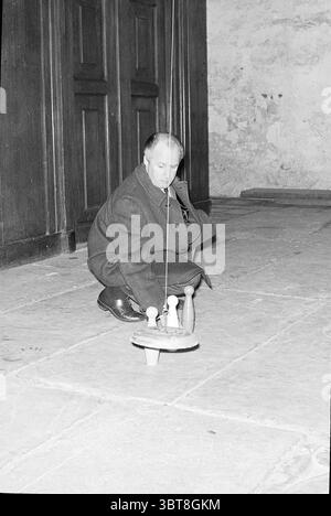 Experiment with sun and earth in Grote Kerk Grote Kerk Haarlem churches of St. Bavokerk Sciences, Whizgle News, Dutch Desk, The Netherlands, 1950 - 2000 on 17-05-1973. The image contains these topics. In a dimly lit room with weathered stone walls, a man is crouched on the floor, intently focused on a unique setup before him. He wears a dark, heavy coat that contrasts sharply with the lighter, worn stone flooring beneath him. The atmosphere feels quiet and contemplative, perhaps suggesting a moment of curiosity or experimentation. Before the man is a circular base, possibly made of wood or a s Stock Photo