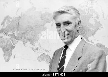 Mr. Otto from ANVR travel agency Reisbureaux, Whizgle News, Dutch Desk, The Netherlands, 1950 - 2000 on 09-12-1985. The image contains these topics. The scene features a man with silver hair styled neatly, presenting a warm and approachable expression. He is dressed in a light-colored suit jacket, complemented by a darker tie, which adds a touch of formality to his appearance. Behind him, a large world map serves as the backdrop, prominently displaying international routes marked by lines traversing various continents. The map has a vintage feel, with muted earth tones, including shades of bei Stock Photo
