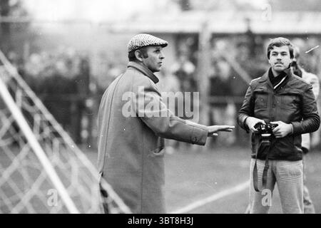 Football match Haarlem - NEC Haarlem The Netherlands, Whizgle News, Dutch Desk, The Netherlands, 1950 - 2000 in 1977. These topics appear in the image. The scene captures a dynamic moment on a sports field, likely during a soccer match. In the foreground, two men are prominently featured. One man, dressed in a long coat with a noticeable checkered cap, gestures emphatically with his left hand, suggesting he is perhaps expressing frustration or giving directions. His serious demeanor and the intensity of his posture convey a sense of urgency or authority. The second man stands beside him, holdi Stock Photo
