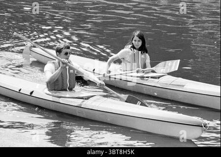 Canoeists in the Leidsevaart Canoe, Whizgle News, Dutch Desk, The Netherlands, 1950 - 2000 on 07-07-1981. The image includes these topics. The scene depicts two individuals navigating in sleek, narrow kayaks gliding across a calm body of water. The setting is tranquil, with softly rippling water reflecting light in gentle patterns, creating a sense of motion and serenity. On the left, a man, wearing dark sunglasses and a light-colored shirt, displays a focused expression as he paddles. His kayak appears streamlined, designed for speed and agility. The oar he’s gripping is wooden, contrasting w Stock Photo