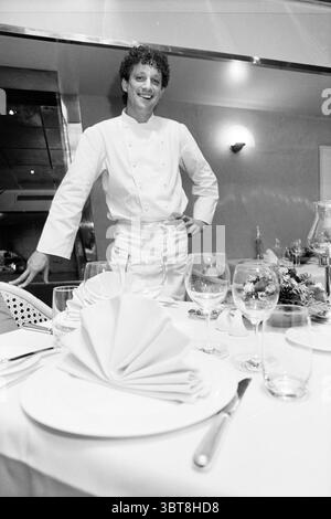 L. Rieve chef at Bokkedoorns restaurant Overveen Overveen, Whizgle News, Dutch Desk, The Netherlands, 1950 - 2000 on 26-11-1996. The image includes these topics. In an upscale dining setting, a cheerful chef stands proudly beside an elegantly arranged table. The atmosphere is warm and inviting, enhanced by soft, ambient lighting that casts a gentle glow across the scene. The chef, wearing a crisp white uniform that gleams under the light, has a relaxed posture with one hand resting on his hip, exuding confidence and friendliness. His hair is curly and dark, adding a touch of vitality to his pr Stock Photo