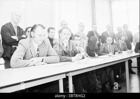 Signing of compromise between strikers and Hoogovens board in the presence of Arie Groenevelt. Velsen-Noord., Whizgle News, Dutch Desk, The Netherlands, 1950 - 2000. The image contains these topics. The scene captures a gathering of men and women in a corporate or formal setting, likely during a meeting or discussion. A long table stretches across the foreground, where a group of men is seated. They exhibit a range of expressions, from concentration to concern. The man in the center is dressed in a light-colored suit, indicating a position of prominence or leadership. To his left, another man Stock Photo