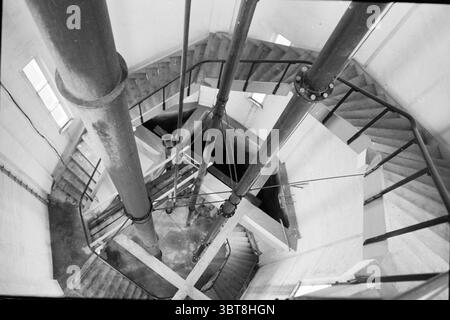 Cleaning inside the water tower Heemstede (spiral staircase) Heemstede The Netherlands, Whizgle News, Dutch Desk, The Netherlands, 1950 - 2000 on 19-09-1994. These are the elements in the image. In the scene, a spiral staircase descends into a well-lit, industrial-looking space. The structure features broad, concrete steps that sweep elegantly downward, creating a sense of movement. The metal handrail, painted black, contrasts sharply with the light-colored concrete walls, which are marked by rough textures. Central to the composition are several large, cylindrical pipes that rise vertically a Stock Photo