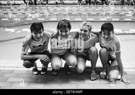 Swimming team HPC (youth) from Heemstede. Swimming Heemstede The Netherlands, Whizgle News, Dutch Desk, The Netherlands, 1950 - 2000 on 06-10-1984. The image shows these topics. In the scene, four young swimmers are posed together, showcasing a cheerful and playful camaraderie. The backdrop features a swimming pool, with lanes marked by floating dividers and clear water reflecting soft lighting. The colors are predominantly cool tones, with deep blues and turquoise from the pool, contrasted by the swimmers' attire. The individuals are dressed in matching white shirts, each adorned with bold le Stock Photo