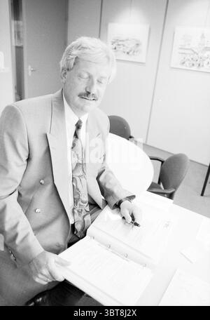 Man behind desk, Whizgle News, Dutch Desk, The Netherlands, 1950 - 2000 on 06-1992. These topics appear in the image. In a professional indoor setting, a man with neatly styled, light-colored hair sits at a round table. He is dressed in a tailored suit that features a light gray jacket with slightly darker gray pants. His tie is patterned, incorporating soft colors that suggest a floral or abstract design. The table is covered with documents that he appears to be reviewing closely. One hand holds a pen, while the other rests on the papers, indicating an engaged and thoughtful demeanor. His exp Stock Photo
