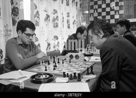 Mr. de Boer HST Blast Furnace Chess Tournament, Whizgle News, Dutch Desk, The Netherlands, 1950 - 2000 on 25-01-1968. These are the elements in the image. In a cozy, well-lit room, a chess match is taking place between two focused individuals sitting across from each other at a wooden table. The players are deeply engrossed in their strategy, displaying an intensity that fills the atmosphere with anticipation and concentration. To the left, a young man with dark, short hair and glasses wears a sweater over a collared shirt. His expression is serious, and he stares intently at the chessboard, a Stock Photo