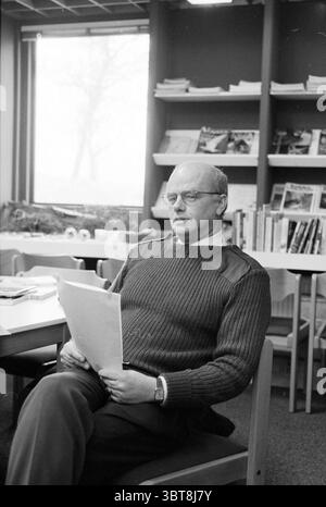 Mr. H. Schouten ranger Kennemerduinen, Whizgle News, Dutch Desk, The Netherlands, 1950 - 2000 on 02-02-1997. These are the elements in the image. In a cozy indoor setting, a man sits comfortably at a wooden table, engaged in reading through a document. His hairstyle is neat, and he wears glasses that lend a scholarly air to his demeanor. He is dressed in a textured, dark sweater with a subtle shine, along with light-colored pants. The surrounding environment showcases a library or study space, with shelves filled with various books and magazines that hint at a rich array of subjects. A large w Stock Photo