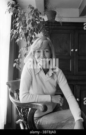 Anitra Kaan Amsterdam Persons, Whizgle News, Dutch Desk, The Netherlands, 1950 - 2000 on 23-06-1977. These are the topics in the image. The scene features a young woman seated comfortably in a wooden chair, exuding a warm, inviting presence. She has shoulder-length blonde hair, styled simply, which complements her friendly smile. The soft, natural lighting pours in from a nearby window, creating a gentle glow that highlights her facial features and the texture of her cozy light-colored sweater. Behind her, a dark wooden cabinet adds depth to the setting, contrasting with the light tones of her Stock Photo