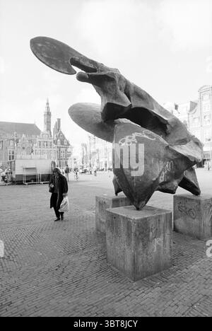 Sunfighter Gr. Market Haarlem Grote Markt The Netherlands, Whizgle News, Dutch Desk, The Netherlands, 1950 - 2000 on 21-03-1991. These are the topics in the image. In a bustling square, a striking abstract sculpture commands attention, composed of jagged, angular forms that seem to twist and curl skyward. The sculpture is crafted from a dark, metallic material, its surface showing hints of patina, which contrasts sharply with the lighter cobblestones beneath it. Nearby, a lone figure ambles through the scene, draped in a long black coat that flows with their movement. They carry a shopping bag Stock Photo