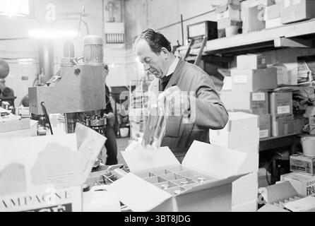 De Pelikaan Distillery Wine, Whizgle News, Dutch Desk, The Netherlands, 1950 - 2000 on 27-12-1978. These topics are shown in the image. The scene unfolds in a bustling workshop filled with an array of wooden shelves stacked with boxes and various tools. A middle-aged man, dressed in a dark apron, is the focal point, engaged in a meticulous task. He grips a bottle in one hand while his other hand hovers over a cardboard box, suggesting an assembly line process. The man has a concentrated expression, indicating a dedication to his work. The color palette is mainly muted, dominated by grays and b Stock Photo
