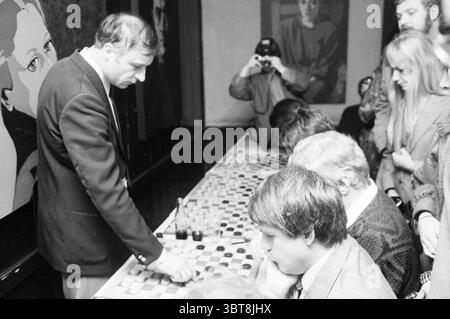 Checkers by Rus in the Halve Maan Santpoort Checkers Santpoort, Whizgle News, Dutch Desk, The Netherlands, 1950 - 2000 on 30-11-1986. These are the elements in the image. The scene captures a lively indoor setting, primarily focused on a long table where several individuals are engaged in a game of checkers. At the forefront, a man in a dark suit leans slightly forward over the board, attentively analyzing his next move. His concentration is palpable, with a furrowed brow that shows his commitment to the game. Surrounding the table are several players, all intently focused on the game. The atm Stock Photo
