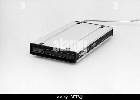 Computer modem Computers, Whizgle News, Dutch Desk, The Netherlands, 1950 - 2000 on 15-01-1987. These are the topics in the image. The scene features a sleek electronic device that rests on a smooth, light-colored surface. The object has a streamlined, rectangular shape with a predominantly silver metallic finish, giving it a modern, minimalist appearance. The edges are subtly rounded, enhancing its contemporary aesthetic. At the front, a small black section contrasts sharply with the silver, housing various buttons and indicators that suggest its functionality. The buttons are arranged in a g Stock Photo