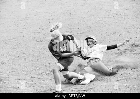 Netherlands - Chicago, Whizgle News, Dutch Desk, The Netherlands, 1950 - 2000 on 16-07-1993. The image shows these topics. In a dynamic outdoor scene, two young athletes engage in an intense moment of a baseball game on a dusty field. The background is a textured sandy surface, scattered with small pebbles and gravel, indicating an active play area. On the left, a player with long, tousled blonde hair rushes toward the base, wearing a loose-fitting, dark-colored shirt and shorts. The player is mid-stride, with a determined expression, arms extended forward as they grasp the base. Stock Photo