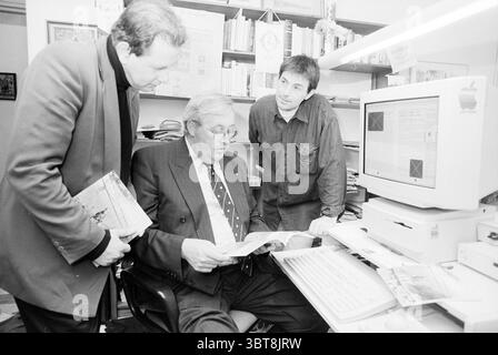 Makers of the book Eye Camp in Nepal, Whizgle News, Dutch Desk, The Netherlands, 1950 - 2000 on 26-10-1994. The image includes these topics. In a well-lit office setting, three individuals are engaged in a discussion. The space is filled with bookshelves that house an array of books, indicating a scholarly atmosphere. A white computer sits prominently on a desk, partially obscured by scattered papers and a keyboard. The man seated at the desk wears a tailored dark suit and is intently examining a document, possibly a report or magazine. His focus suggests he holds a position of authority or ex Stock Photo