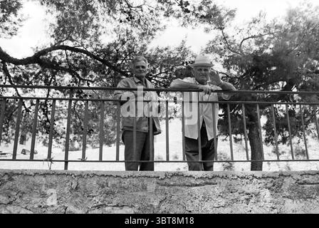 Travel to Mallorca Travel, Whizgle News, Dutch Desk, The Netherlands, 1950 - 2000 on 22-08-1979. These are the elements in the image. In a serene outdoor setting, two elderly men stand beside a low stone wall, leaning casually against a railing. The background is filled with lush greenery, primarily trees with broad leaves that provide dappled shade. The environment gives off a tranquil, natural ambiance, enhanced by the soft interplay of light filtering through the foliage. The men are wearing simple yet distinct outfits. One man, dressed in a light-colored shirt, appears relaxed, with a thou Stock Photo