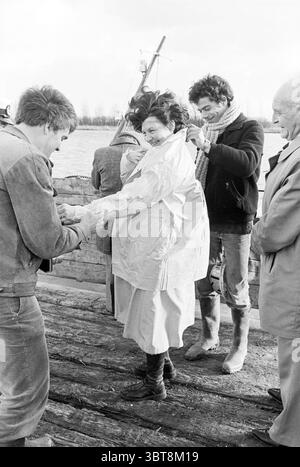 First pole marina sailing club in side channel C First pole Sailing IJmuiden The Netherlands, Whizgle News, Dutch Desk, The Netherlands, 1950 - 2000 on 10-12-1979. These topics are shown in the image. In a rustic setting by the water, several individuals gather on a weathered wooden dock. The backdrop features a calm, reflective body of water, framed by a slightly overcast sky, hinting at a chilly atmosphere. The scene is infused with a sense of camaraderie and light-heartedness. Prominently, a woman stands in the center, wearing a long, light-colored coat that billows gently in the breeze. He Stock Photo