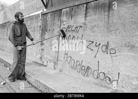 Defaced wall at HFC Haarlem Muren, Whizgle News, Dutch Desk, The Netherlands, 1950 - 2000 on 08-11-1976. These are the elements in the image. The scene depicts a gritty urban setting, marked by a concrete wall that serves as a canvas for graffiti. The wall is weathered, with a rough texture that emphasizes its age. Bold, dark graffiti scrawls are present, contrasting sharply against the light grey of the concrete. A solitary figure stands in the foreground, focused on actively cleaning the wall. This person is dressed in a dark, cozy sweater that suggests a casual, working-class vibe, with loo Stock Photo